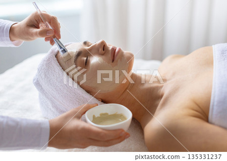 Therapist gently spreading cosmetic clay mask with brush to woman at spa Therapist gently spreading cosmetic clay mask with brush to woman at spa 135331237