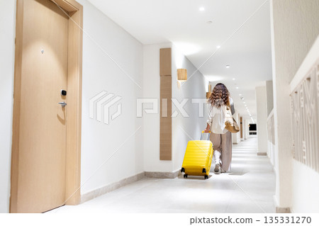 Rear view of adult caucasian woman walking down hotel corridor pulling suitcase 135331270