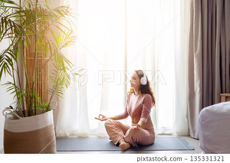 Adult caucasian woman meditating with headphones on yoga mat in hotel room 135331321