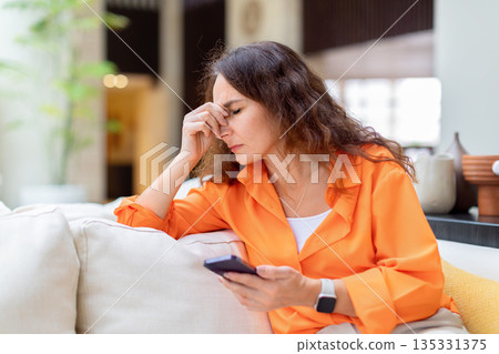 Stressed woman holding smartphone and touching face while sitting in hotel lobby 135331375