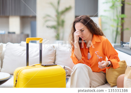 Stressed woman holding glasses and touching face while sitting in hotel lobby 135331386