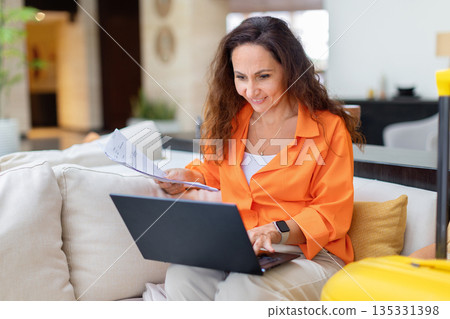 Adult woman working with laptop and documents while sitting in hotel lobby Adult woman working with laptop and documents while sitting in hotel lobby 135331398