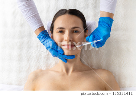 Front view of young woman smiling gently during professional cosmetic facial injection 135331411