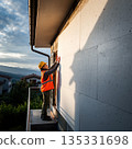 Construction worker installs material on building wall during sunset near residential area with mountains in background 135331698