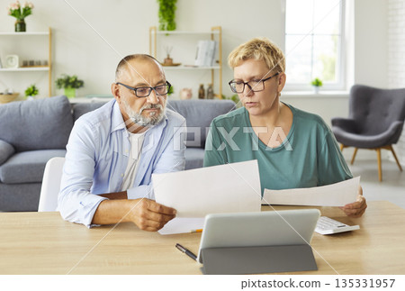 Senior couple reviewing bills and finances at home with tablet and papers 135331957