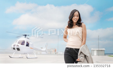 Young Asian woman in luxury lifestyle standing with a private helicopter on a helipad. Concept of executive travel, high-end tourism, aviation business, and success Young Asian woman in luxury lifestyle standing with a private helicopter on a helipad. Concept of executive travel, high-end tourism, aviation business, and success 135332718