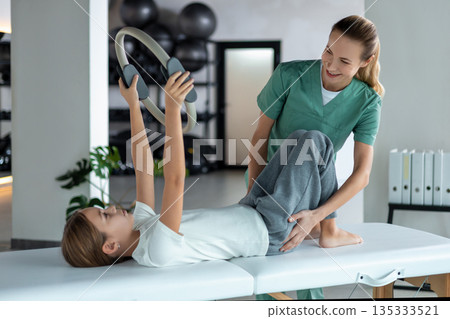 Pediatric physiotherapist assisting girl with Pilates ring exercise Pediatric physiotherapist assisting girl with Pilates ring exercise 135333521