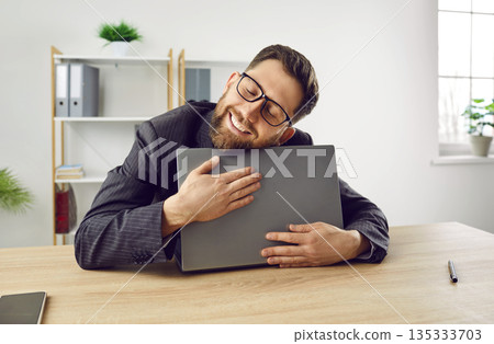 Funny happy young businessman or office worker loves and hugs his laptop computer Funny happy young businessman or office worker loves and hugs his laptop computer 135333703