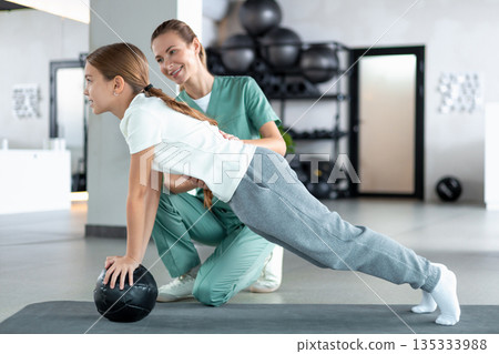 Female therapist helping little girl holding plank on mat strengthening muscle Female therapist helping little girl holding plank on mat strengthening muscle 135333988