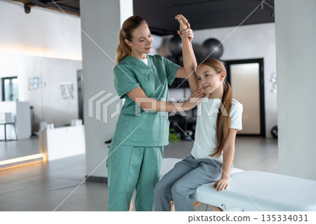 Woman therapist helping little girl stretching arm sitting on couch 135334031