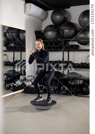 Blonde young woman in black sportswear exercising on a balance board 135334179