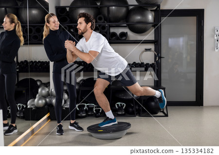 Man having a workout on a balance board, female coach helping him 135334182