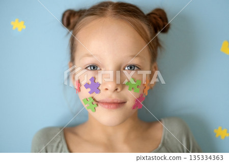 Portrait of a girl with pieces of multicolored puzzles pasted on her cheeks, a symbol of autism. Support and care for people with autism spectrum disorder. Education and social equality. Portrait of a girl with pieces of multicolored puzzles pasted on her cheeks, a symbol of autism. Support and care for people with autism spectrum disorder. Education and social equality. 135334363