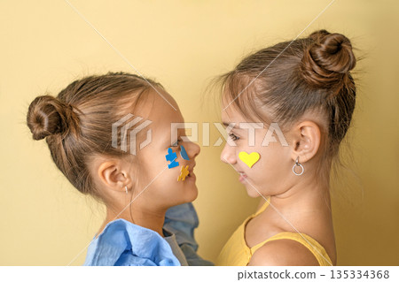 Two cheerful cheerful girls on a yellow studio background with decorations on their faces in the form of blue and blue puzzles. World Down Syndrome Day. 135334368