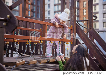 Mother and child playing on a playground bridge, holding hands and smiling 135334423