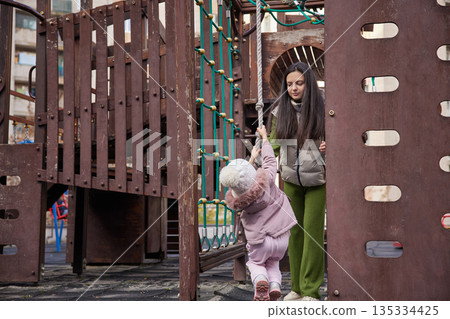 Mother assisting young child climbing rope on wooden playground structure 135334425