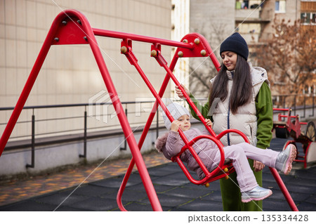 mother pushing daughter on a swing at outdoor playground 135334428
