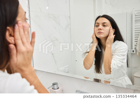 woman applying skincare cream to her face in white bathroom mirror performing a beauty routine 135334464