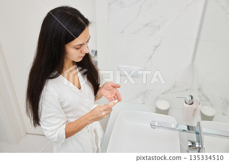 Woman in bathrobe applying moisturizing cream to her hand over a sink 135334510