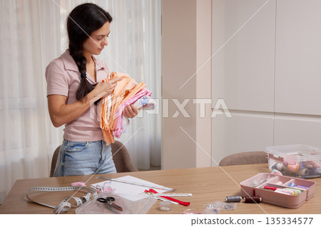 Woman holding various colorful fabrics, organizing her needlework tools on a table for a craft project 135334577