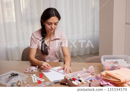 Woman focusing on drawing a fashion sketch at a table with sewing supplies Woman focusing on drawing a fashion sketch at a table with sewing supplies 135334579