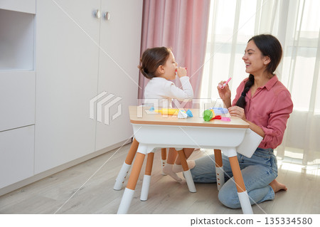 Mother and daughter celebrating and playing with party blowers at a small table 135334580