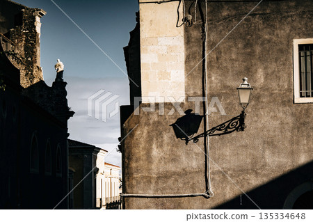 Light And Shadow On Catania Street Wall: Italian Sicilian Urban Scene With Weathered Facade, Window, And Street Lamp In Strong Sunlight 135334648