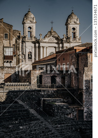 Teatro Romano E Odeon In Catania, Sicily: Ancient Roman Theater Ruins With Stone Seating, Historic Buildings, And Church Towers Under Clear Daylight 135334671