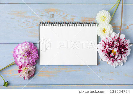 Notebook with clean page with purple flowers dahlias on blue wooden background. Colorful flower background with space for text. Nature trendy decorative design. Flat lay. Notebook with clean page with purple flowers dahlias on blue wooden background. Colorful flower background with space for text. Nature trendy decorative design. Flat lay. 135334734
