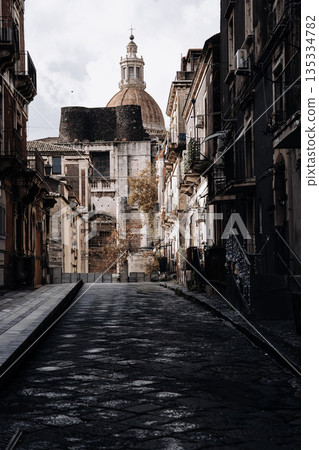 Narrow Street In Catania With Church Dome: Italian Sicilian City Scene With Historic Stone Buildings, Strong Light Contrast, And Deep Urban Perspective Narrow Street In Catania With Church Dome: Italian Sicilian City Scene With Historic Stone Buildings, Strong Light Contrast, And Deep Urban Perspective 135334782
