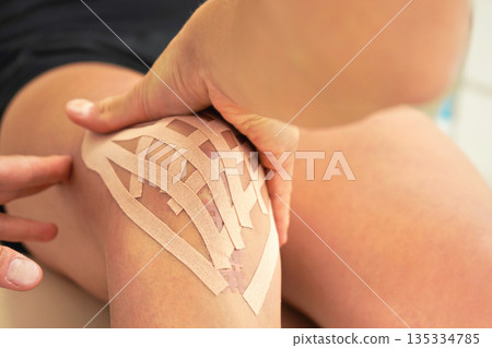 Young female physiotherapist applying kinesiology tape and massaging knee of her patient, closeup detail on hands Young female physiotherapist applying kinesiology tape and massaging knee of her patient, closeup detail on hands 135334785