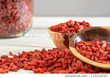 Dried goji berries in wooden bowls, scattered over white boards table under, blurred large glass jar full of fruits in background 135334797