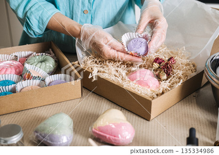 Person wearing protective gloves packs handmade purple bath bomb into gift box filled with shredded paper. Artisanal business, home-based startup, craft industry 135334986