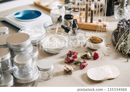 Various natural cosmetic ingredients botanical powders, dried flowers, and essential oils sit on workbench beside digital scale and glass jars. Sustainable beauty, clean skincare, organic formulation. 135334988