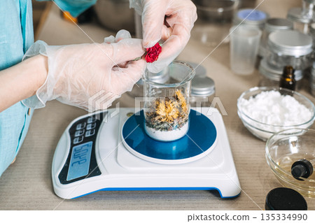 Person in protective gloves adds dried flower to glass beaker on electronic scale containing botanical ingredients. Botanical extracts, natural formulation, plant-based ingredients. 135334990