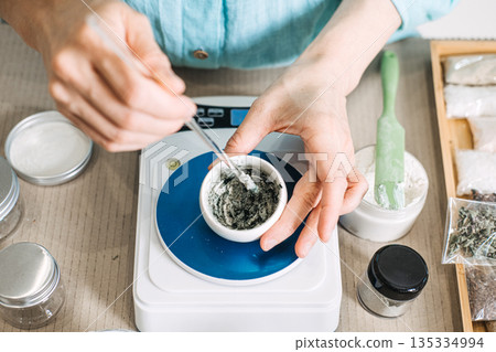 Hands mix green paste in small bowl on digital scale surrounded by laboratory equipment and jars. Small business start-up, handmade skincare, artisanal beauty products, home-based laboratory. Hands mix green paste in small bowl on digital scale surrounded by laboratory equipment and jars. Small business start-up, handmade skincare, artisanal beauty products, home-based laboratory. 135334994