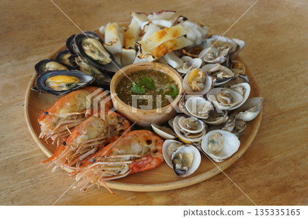 Closeup Grilled seafood, three large shrimps, cut squid, cockles, scallops and seafood dipping sauce in a wooden bowl on a wooden tray, on a wooden table background, food, delicious, hot, copy space 135335165