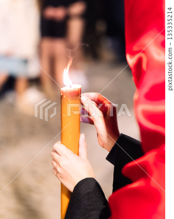 Close up of a person in a red hood holding a lit wax candle at night. Close up of a person in a red hood holding a lit wax candle at night. 135335214