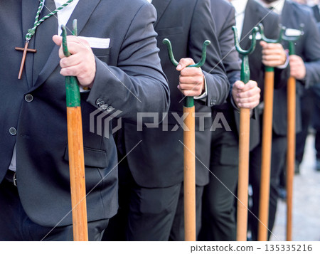Men in black suits carrying traditional metal staffs during a religious procession 135335216