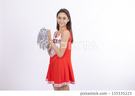 Young cheerleader girl holding silver pom poms near her smiling face in studio 135335225