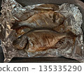 Two whole sea bream baked in foil with crispy skin and light seasoning resting on a roasting tray in warm kitchen light 135335290