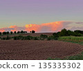 Wide rural landscape with plowed fields and distant trees under glowing pink evening clouds. Calm countryside scene with soft sky colors and open horizon 135335302