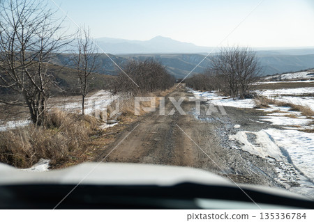 Rural dirt road in Armenia with melting snow, bare trees, rolling hills, distant mountains under winter light, creating sense of journey and openness Rural dirt road in Armenia with melting snow, bare trees, rolling hills, distant mountains under winter light, creating sense of journey and openness 135336784