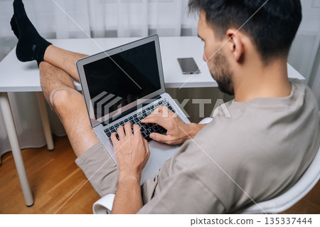 Young man working remotely on laptop in cozy home office, feet up on desk, casual t-shirt and shorts, focused and relaxed while handling freelance work and calls. Young man working remotely on laptop in cozy home office, feet up on desk, casual t-shirt and shorts, focused and relaxed while handling freelance work and calls. 135337444
