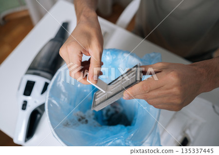 Person cleaning HEPA filter from dirty robot vacuum cleaner, carefully tapping out accumulated dust and debris into bin with blue plastic liner, performing essential device maintenance. Person cleaning HEPA filter from dirty robot vacuum cleaner, carefully tapping out accumulated dust and debris into bin with blue plastic liner, performing essential device maintenance. 135337445