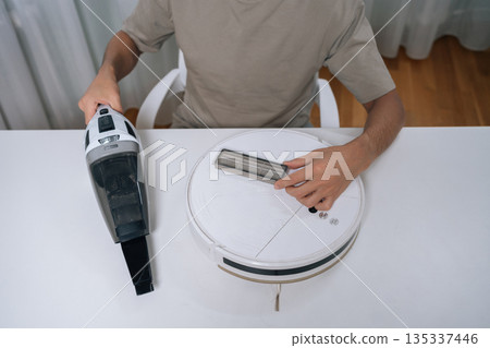Top view of person performing routine maintenance on robot vacuum cleaner, using handheld vacuum to clean dust filter for optimal smart home appliance performance and longevity, close-up cropped shot. Top view of person performing routine maintenance on robot vacuum cleaner, using handheld vacuum to clean dust filter for optimal smart home appliance performance and longevity, close-up cropped shot. 135337446