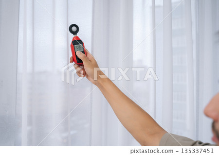 Close-up cropped shot of man measuring natural light near window with digital luxmeter, testing illumination levels for optimal home brightness, energy efficiency and comfortable indoor daylighting. 135337451
