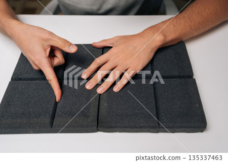 Man testing soundproof acoustic foam panels with fingers, checking texture and material density for sound absorption qualities in a controlled environment, close-up cropped shot. 135337463