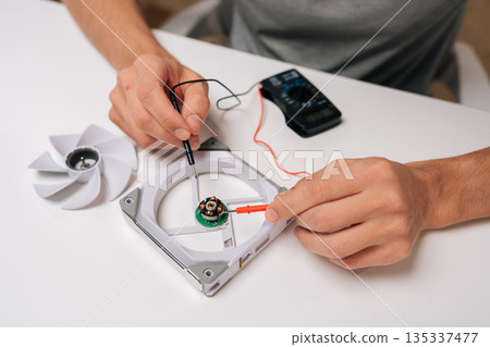 Top view of electric troubleshooting and checking disassembled computer cooling fan motor with digital multimeter on white desk, focusing on electronics and pc hardware repair, close-up cropped shot. 135337477
