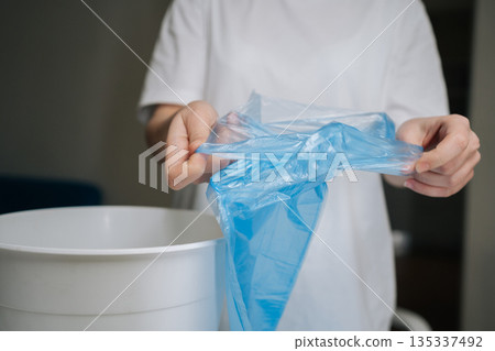 Hands unfolding and positioning fresh blue plastic bag into empty white trash can, signifying start of efficient waste management and cleanliness routine in domestic setting, close-up cropped shot. Hands unfolding and positioning fresh blue plastic bag into empty white trash can, signifying start of efficient waste management and cleanliness routine in domestic setting, close-up cropped shot. 135337492